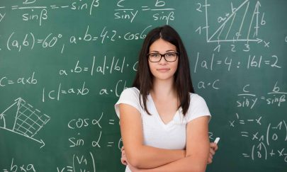 Intelligent young female maths student or teacher wearing glasses standing in front of a chalkboard with mathematical equations with folded arms and a piece of chalk in her hand