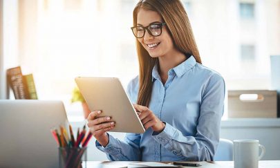 Lets check my timetable! Cheerful young beautiful woman in glasses using her touchpad with smile while sitting at her working place