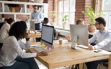 Multi-ethnic employees working on computers in modern office room, corporate staff team people diverse workers group using desktop monitors sitting at desks in open shared coworking space together