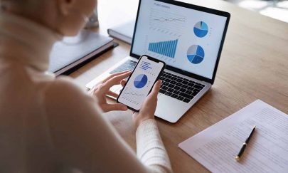 Woman hold smartphone use pc at workplace. Project stats financial data sales charts on laptop and cellphone screen, close up view over shoulder. Report preparation, synchronization for safety concept